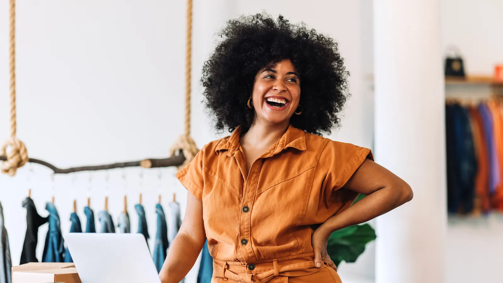 A smiling small business owner stands at a laptop in her clothing shop.